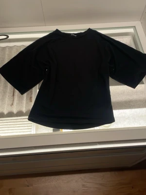 Svart t-shirt från 157 med vida ärmar - Svart t-shirt från 157 i storlek XS. Modellen har breda, korta ärmar och en klassisk rund halsringning. T-shirten är enkel och stilren, perfekt för dig som gillar minimalistisk look. Materialet känns mjukt och bekvämt mot huden.