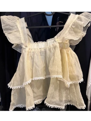 Beige volangig linnetopp med spetsdetaljer - Supersöt beige linnetopp med volanger och breda axelband. Toppen har två lager med volanger och små vita spetsbollar längs kanterna. Tunt, luftigt tyg som ger en romantisk vibe – perfekt för varma dagar. Volangärmarna är avtagbara om man vill ha off shoulder look!!!