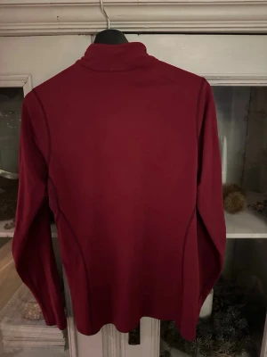 Vinröd underställ från Arc'teryx - Säljer ett vinrött underställ från Arc'teryx med lång ärm och half zip framtill. Toppen har ett stilrent Arc'teryx-tryck i gult på ärmen och är tillverkad i ett mjukt syntetmaterial som passar perfekt för träning eller utomhusaktiviteter.