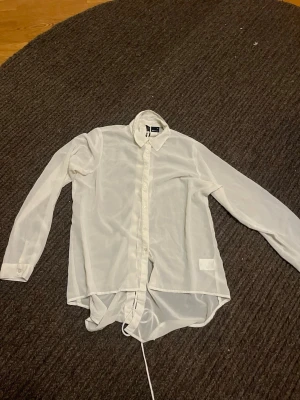 Vit skjorta med snörning från Gina Tricot - Säljer en vit, transparent skjorta från Gina Tricot i storlek 38. Skjortan har lång ärm, klassisk krage och cool snörning längs hela ryggen. Materialet är lätt och känns luftigt, perfekt för dig som gillar att sticka ut med detaljer. Knappar framtill och rundad nederkant.