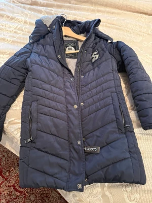 Mörkblå quiltad pufferjacka Staccato - Säljer en mörkblå pufferjacka från Staccato med quiltad design och mjukt grått fleecefoder. Jackan har huva, dragkedja och knappar framtill samt praktiska fickor med dragkedja. Perfekt för kalla dagar och riktigt skön att ha på sig.