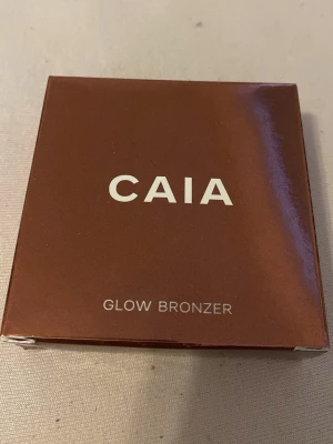 CAIA Glow Bronzer La Digue - Snygg bronzer från CAIA i nyansen La Digue. Kommer i en stilren, brun metallisk dosa med vitt CAIA-tryck. Innehåller 10 g och ger ett glowigt, solkysst resultat. Perfekt för att framhäva ansiktskonturer och skapa värme i looken. Den är i nytt skick, min mamma köpte fel färg till mig i julklapp tyvärr så därför säljer jag:)