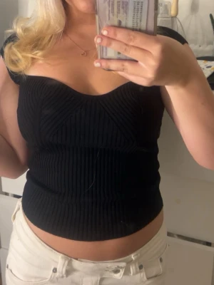 Svart ribbad korsett-topp - Säljer en svart korsett-topp med ribbad struktur och snygg sweetheart-urringning. Toppen är tight och har korta ärmar som sitter precis på axlarna. Perfekt att styla med jeans eller kjol för en trendig look.