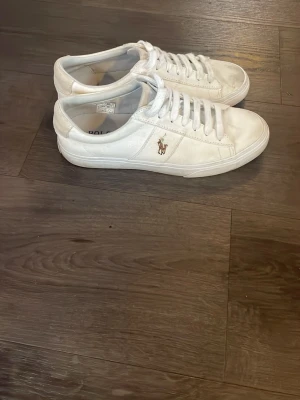 Vita sneakers Polo Ralph Lauren EU 42 - Snygga vita sneakers från Polo Ralph Lauren med klassisk logga på sidan. Skorna har rund tå, snörning och platt sula i svart. Tillverkade i tyg med stilren design och boxen medföljer. Perfekt för dig som gillar clean och tidlös stil. Dom är andvända fåtal gånger 