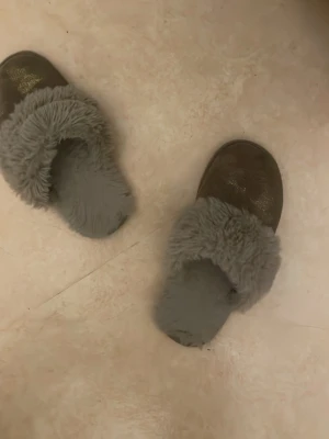 Bruna fluffiga tofflor med päls - Mysiga bruna tofflor med grå fluffig päls på insidan och ovansidan. Perfekta för att hålla fötterna varma hemma under kyliga dagar. Tofflorna har en rund tå och platt sula för extra komfort.