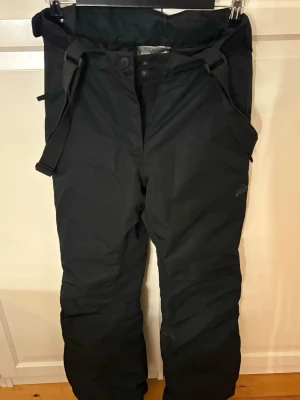 Svarta överdragsbyxor från McKinley  - Säljer ett par svarta täckbyxor från McKinley i storlek 36. Byxorna har justerbara hängslen, hög midja och stängs med både dragkedja och knappar. Perfekta för vinterns alla äventyr och håller dig varm och torr i backen. Köp nu jag postar snabbt!!💗💕