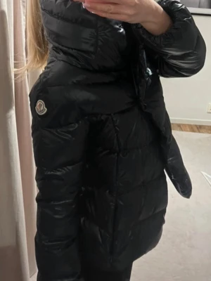 Svart dunjacka från Moncler - Säljer en svart, glansig dunjacka från Moncler med hög krage och quiltad design. Jackan har lång ärm, knappar framtill och klassisk Moncler-logga på ena ärmen. Sååå fin i nyskick
