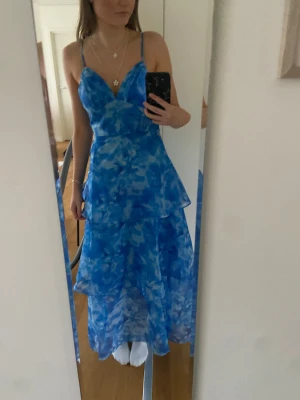 Blommig blå långklänning med volanger - Säljer en superfin blå långklänning med blommigt mönster och volanger i flera lager. Klänningen har smala axelband och v-ringad urringning.