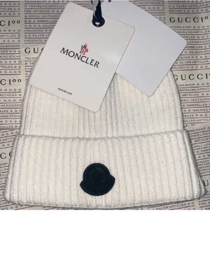 Vit ribbad mössa från Moncler - Säljer en snygg vit ribbad mössa från Moncler med svart logotyp framtill. Mössan har en klassisk uppvikt kant och är tillverkad i mjukt material som håller dig varm. Perfekt för kyliga dagar och enkel att matcha med din vinterstil.