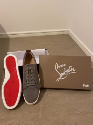 Grå sneakers från Christian Louboutin - Säljer ett par gråa sneakers från Christian Louboutin med klassisk röd sula och vit kant. Skorna har rund tå, snörning och är tillverkade i mocka med snygga detaljer. Kommer med originalkartong, dustbag och äkthetskort från Paris. KAN PRUTAS FÖR SERIÖSA KÖPARE.