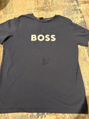 Blå BOSS t-shirt med tryck - Snygg marinblå t-shirt från BOSS med vit BOSS-tryck på bröstet. Modellen är regular fit och har rund hals samt korta ärmar. Tillverkad i mjuk bomull med solskydd och ansvarsfullt framtaget material. Perfekt för en chill och stilren look.