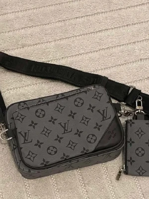  axelväska i grått - Säljer en snygg axelväska  i grått med klassiskt monogrammönster. Väskan har en bred svart axelrem och en mindre matchande plånbok som hänger på sidan. Materialet är skinn och väskan har dragkedja upptill.