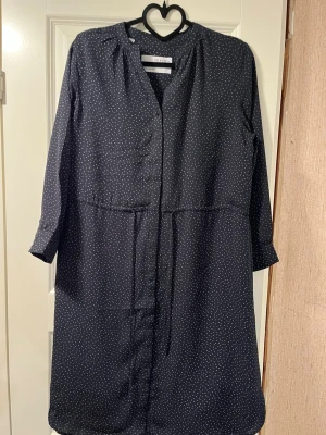 Mörkblå prickig klänning Selected Femme - Mörkblå knälång klänning från Selected Femme med små vita prickar, rak passform med dragsko i midjan och  trekvartsärmar. Klänningen har enkel knäppning framtill och ett tunt, luftigt material som känns skönt mot huden. Perfekt för dig som gillar stilrena mönster och en avslappnad look.