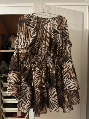 Zebramönstrad volangklänning - Säljer en kort klänning med zebramönster i beige, svart och bruna toner. Klänningen har volanger, resår i midjan och vida ärmar. Materialet är lätt och luftigt, perfekt för dig som vill sticka ut med en cool animal print. Aldrig använt med prislapp