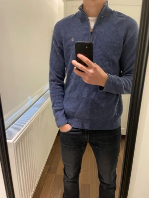 Blå stickad half zip-tröja Morris - Säljer en blå stickad tröja från Morris med half zip och liten broderad logga på bröstet. Tröjan har ribbad krage, muddar och är långärmad. Perfekt för dig som gillar klassisk stil med en modern touch. Den är lite skrynklig på bilderna då den har blivit använd på ett tag 