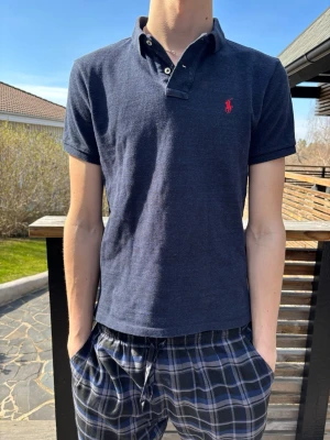 Mörkblå pikétröja från Ralph Lauren - Snygg mörkblå pikétröja från Ralph Lauren med klassisk krage och korta ärmar. Tröjan har en röd broderad logga på bröstet och knappar vid halsen. Piken är i bra skick och sparsamt använd