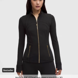 Svart zip hoodie med guld-detaljer - Snygg svart hoodie med guldiga dragkedjor framtill och vid fickorna. Figurnära passform och långa ärmar med tumhål. Perfekt för träning eller chill. Stilren och sportig look.