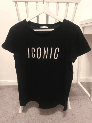 Svart t-shirt med tryck - Svart t-shirt med vitt tryck 'ICONIC' på bröstet. Klassisk rund hals och korta ärmar, perfekt för en chill och stilren look. Materialet känns mjukt och skönt mot huden, och passformen är normal. Enkelt att matcha med jeans eller shorts.