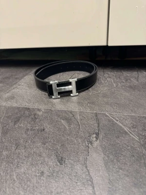 Svart skärp med H-spänne - Snyggt svart skärp med ett stort H-format metallspänne i silverfärg. Skärpet har vit kontrastsöm längs kanterna och är gjort i ett strukturerat skinnmaterial. Perfekt för att ge outfiten en extra detalj och passar till både jeans och kostymbyxor.