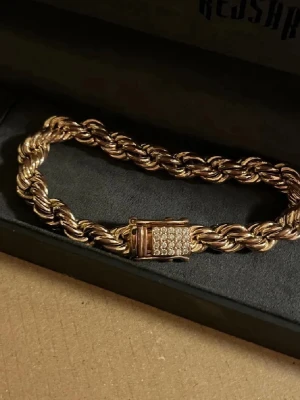 Ingen väst på bilderna - Bilderna visar ett armband i guldton med tvinnad kedja och lås dekorerat med små stenar, ingen väst finns med. Box ingår inte