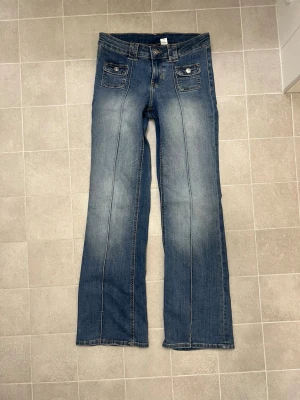 Blå wide jeans från H&M Divided - Snygga blå jeans från H&M Divided med breda ben och raka sömmar framtill. Storlek 38, hyfsat lågmidjade. Modellen har två fickor med lock och knapp framtill, klassisk knappstängning och bälteshällor.