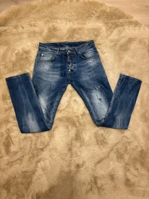  jeans från Dsquared2 - Säljer ett par blå  jeans från Dsquared2 med slitningar och färgstänk för en cool, edgy look. Jeansen har fem fickor, knappgylf och klassisk Dsquared2-lapp bak i midjan. Materialet är jeans med en tvättad finish och detaljerade slitningar. Kom dm för eventuella frågor 🧐pris kan diskuteras 💯😊