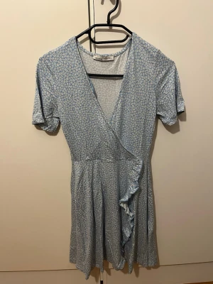 Blommig omlottklänning från Pull&Bear - Söt kort omlottklänning från Pull&Bear i ljusblått med små vita blommor. Klänningen har v-ringning, korta ärmar och volangdetalj längs omlottkanten. Den är endast använt ett fåtal gånger och säljer pågrund av att storleken inte passar 