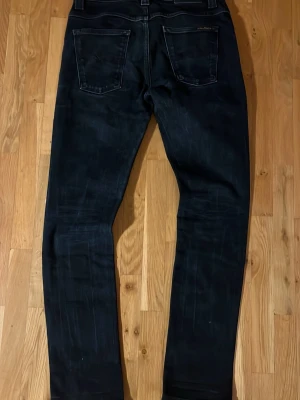 Nudie Jeans Grim Tim  - Säljer ett par svarta Grim Tim Black Ring jeans från Nudie Jeans i storlek 32. Jeansen är tillverkade av 99% ekologisk bomull och 1% elastan för lite stretch. Klassisk femficksmodell med raka ben och diskreta svarta sömmar på bakfickorna.