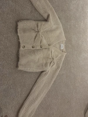 Beige stickad kofta från Zara - Säljer en beige, stickad kofta från Zara med ribbad struktur och snygga rosettdetaljer framtill. Koftan har v-ringning, knappar och långa ärmar. Perfekt för dig som gillar mjuka och mysiga plagg med lite extra stil.