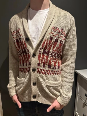 Beige kofta från Hollister med mönster - Säljer en beige stickad kofta från Hollister i storlek M. Koftan har snyggt rödbrunt och vinrött mönster på fram- och baksidan, knappar framtill och två fickor. Perfekt för höst och vinter när man vill vara både varm och stilren.