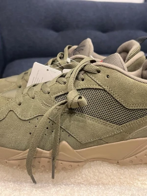 Nike Air Jordan 92 MVP sneakers olivgrön - Nike Air Jordan 92 MVP sneakers i olivgrön mocka med meshpaneler på sidorna och beige sula. Skorna har snörning, svart Nike Air-logga på hälen och röd Jumpman-detalj på plösen och innersulan. Perfekt för dig som gillar streetwear och snygga detaljer.