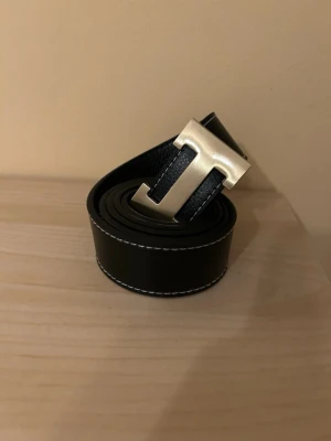 Ingen väst på bilden - Bilden visar ett svart bälte med en stor metallspänne i form av bokstaven H, och vita sömmar längs kanterna. Ingen väst syns på bilden.