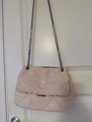 Beige axelväska med kedja - En beige axelväska med fluffigt material som ger en mysig vibe. Väskan har en silverfärgad kedja som axelrem och ett litet metallspänne framtill. Perfekt för dig som gillar trendiga accessoarer med lite extra textur.