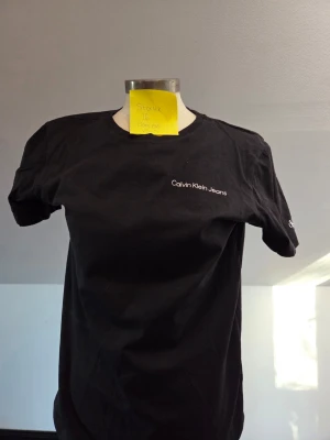 Svart t-shirt från Calvin Klein Jeans - Svart t-shirt från Calvin Klein Jeans med vit logotyp på bröstet och liten CK-detalj på ärmen. Klassisk rund hals och korta ärmar. Tillverkad i mjuk bomull som känns skön mot huden. Perfekt för en clean och enkel look.