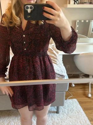 Svart och röd leopardmönstrad klänning från Zara - Säljer en kort klänning från Zara med svart botten och rött leopardmönster. Klänningen har långärmade transparenta ärmar, v-ringning med knappar framtill och smock i midjan. Volangdetaljer vid halsen och ärmslut ger en extra touch. Perfekt för dig som gillar trendiga mönster.❤️