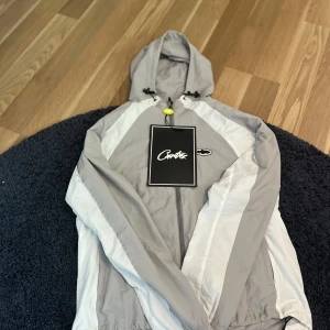 Grå och vit vindjacka från Corteiz - Säljer en stilren grå och vit vindjacka från Corteiz med huva och dragkedja. Jackan har meshfoder, justerbar dragsko i huvan och snygga vita paneler längs ärmarna. Perfekt för dig som gillar streetwear och vill ha en lätt jacka med cool look.