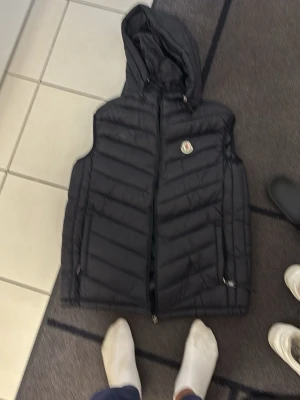 Svart dunväst från Moncler med huva - Säljer en svart dunväst från Moncler med huva och dragkedja framtill. Västen har ett quiltat mönster och två fickor med dragkedja. Moncler-loggan sitter på bröstet och västen har en snygg, sportig look som passar perfekt till lager-på-lager-stil.
