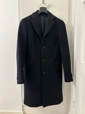 Acne Studios svart wool coat storlek 50 - Klassisk svart rock från Acne Studios. Rocken har en stilren design med tre knappar framtill och fyra knappar vid ärmsluten. Fickor finns på sidorna och på insidan. Materialet är ull och ger en stilren look som funkar till många outfits. Perfekt för dig som gillar enkel och tidlös stil.