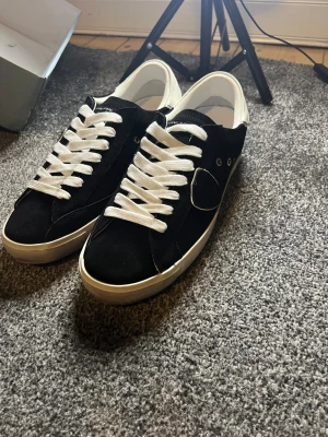 Svarta Philippe Model sneakers - Snygga svarta sneakers från Philippe Model med vit häl och vita snören. Skorna har en rund tå, platt sula och är tillverkade i mocka och skinn. Klassisk design med diskret logga på sidan och detaljer i vitt. Perfekta för dig som gillar stilrena sneakers.