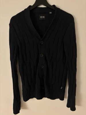 Mörkblå kofta från Only & Sons - Snygg mörkblå kofta från Only & Sons med kabelstickat mönster och fem knappar framtill. Koftan har långärmad passform och ribbade muddar vid ärmslut och nederkant. Perfekt att slänga på över en t-shirt eller hoodie för en chill look.