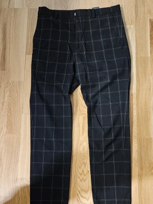Svarta rutig kostymbyxor H&M slim fit - Svarta kostymbyxor från H&M i slim fit-modell med vitt rutmönster. Byxorna har dragkedja, knappstängning och bälteshällor. Materialet är mjukt och lätt, perfekt för en clean och stilren look. Passformen är lite pösigare även om det står slimfit. midjan är normal.
