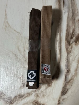 Bruna karate bälten  - Bilderna visar två bruna bälten från Budo-Nord, inte skor. Bältena är tillverkade av bomull och polyester och är 300 cm långa. Märkena och detaljerna indikerar att de är avsedda för kampsport.