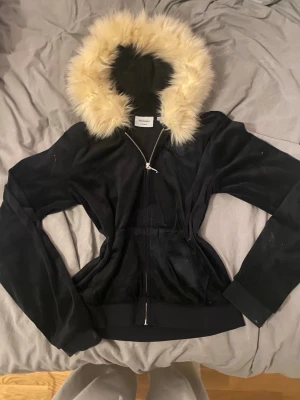 Svart teddyjacka från Juicy Couture - Svart zip up från Juicy Couture med fluffig beige fuskpäls runt huvan. Jackan har dragkedja framtill och är i ett mjukt, syntetmaterial som känns lyxigt. Perfekt för dig som vill ha en mysig och snygg jacka med ikonisk 2000-talsvibe. Passar s-m