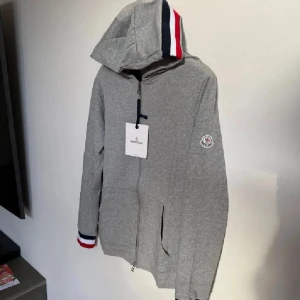 Moncler hoodie - Säljer en stilren grå hoodie från Moncler med dragkedja och randiga detaljer i rött, vitt och blått på huva och ärmslut. Perfekt för en casual look. 🩶(storlek M). Hoodien är äkta också.