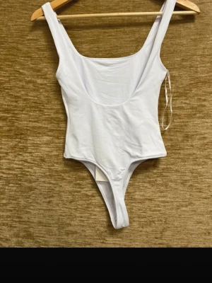 Vit ärmlös bodysuit  - Säljer en vit, ärmlös bodysuit i storlek S med breda axelband och djup rundad halsringning. Modellen är tight och har en enkel, stilren design utan mönster eller detaljer. Materialet känns mjukt och stretchigt, perfekt för att styla med jeans eller kjol.
