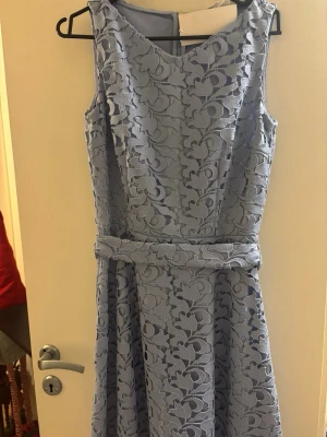 Ljusblå spetsklänning från Hugo Boss - Superfin ljusblå klänning från Hugo Boss med blommigt spetsmönster och rund hals. Klänningen är ärmlös och har en markerad midja med ett matchande bälte. Materialet är spets med polyesterfoder och dragkedja bak. Perfekt för dig som gillar eleganta detaljer.
