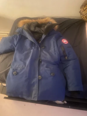 Blå parkajacka från Canada Goose - Säljer en blå parkajacka från Canada Goose med päls på huvan och klassisk logga på ärmen. Jackan har flera fickor med knappar och dragkedja på ärmen. Perfekt för kalla dagar och har en snygg, robust look.