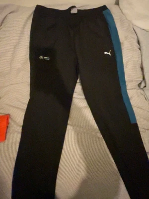 Svarta Puma träningsbyxor med blå detalj - Svarta träningsbyxor från Puma med en blå sidopanel längs benet och Mercedes AMG Petronas Formula One Team-logga. Byxorna har smal passform och är gjorda i polyester, perfekt för sport och träning. Puma-loggan syns tydligt på framsidan.
