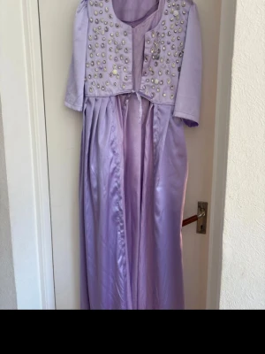kurdisk klänning - Säljer en elegant ljuslila långklänning med glansigt tyg och korta ärmar. Överdelen har vackra pärldetaljer och broderade mönster, samt ett knytband i midjan. Klänningen har en mjuk, flytande siluett och passar perfekt för speciella tillfällen.