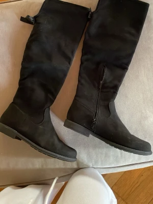 Svarta höga stövlar med dragkedja - Säljer ett par svarta höga stövlar med platt sula och rund tå. Stövlarna har en snygg dragkedja på sidan och är tillverkade i ett mockaliknande material. Perfekta för dig som gillar stilrena och klassiska boots med enkel design.